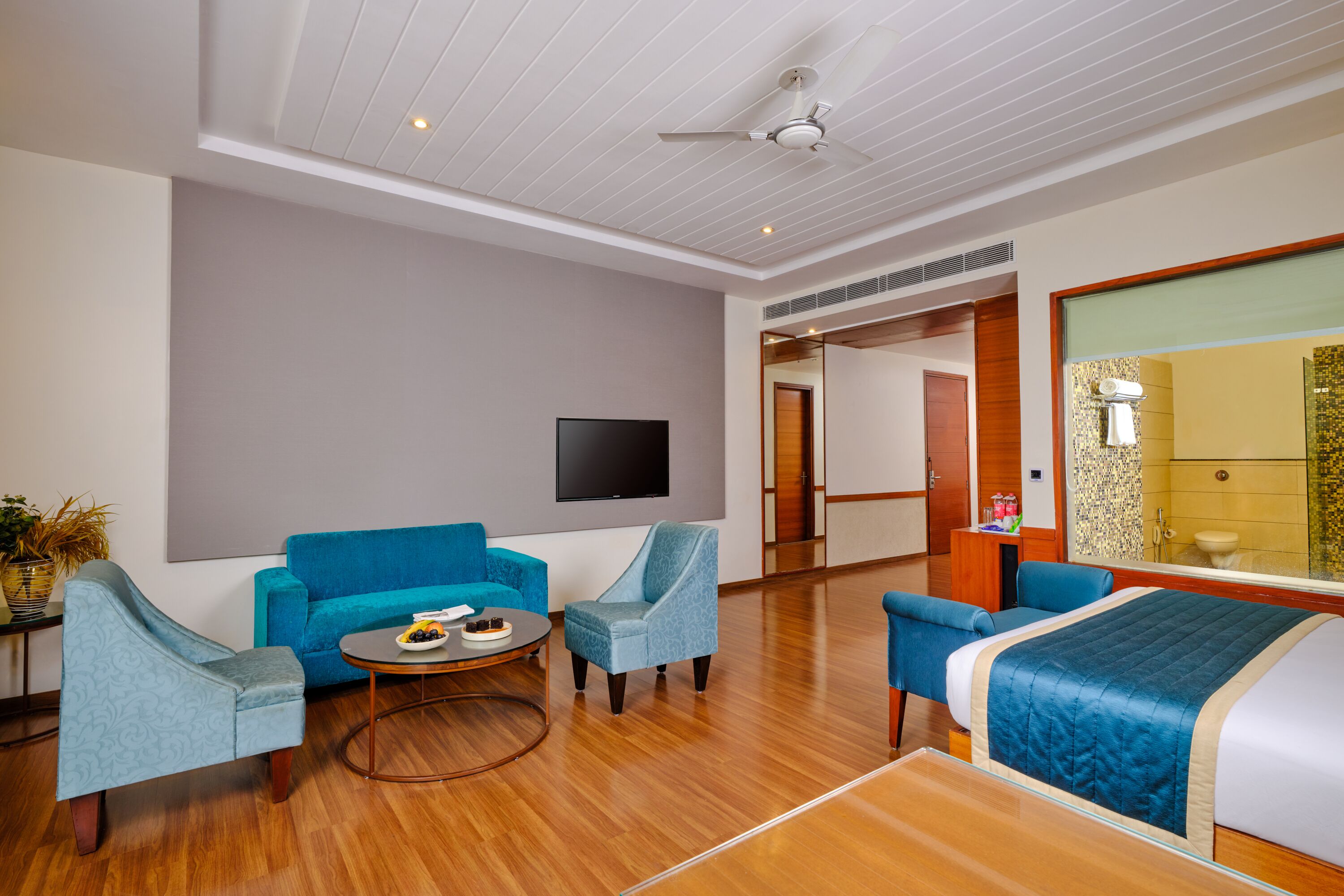 A modern hotel room with a large bed, two armchairs, a sofa, a flat-screen TV, and a wooden floor.