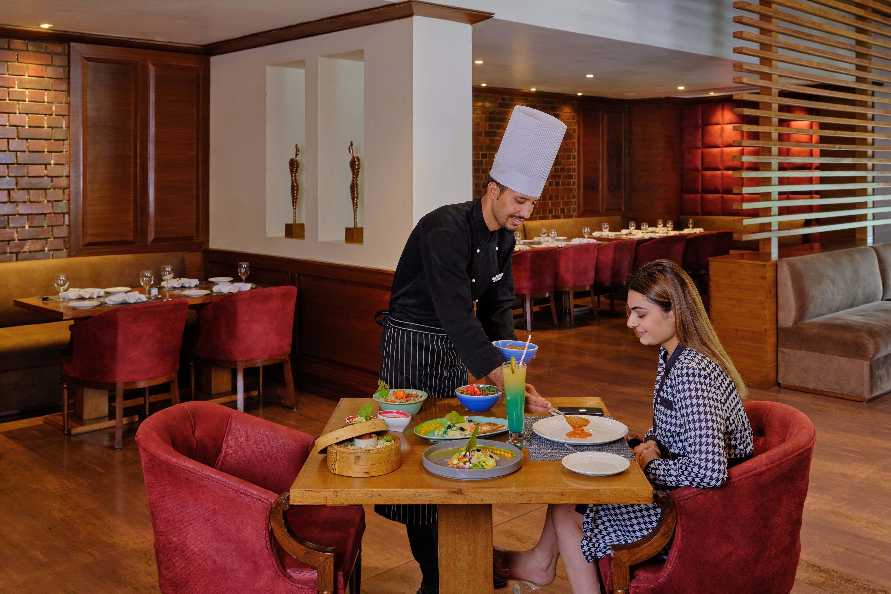 Un restaurante con mesas de madera y sillas tapizadas en rojo, un chef sirviendo comida y una mujer cenando.