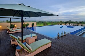 A rooftop infinity pool with lounge chairs and an umbrella, surrounded by a wooden deck and potted plants.