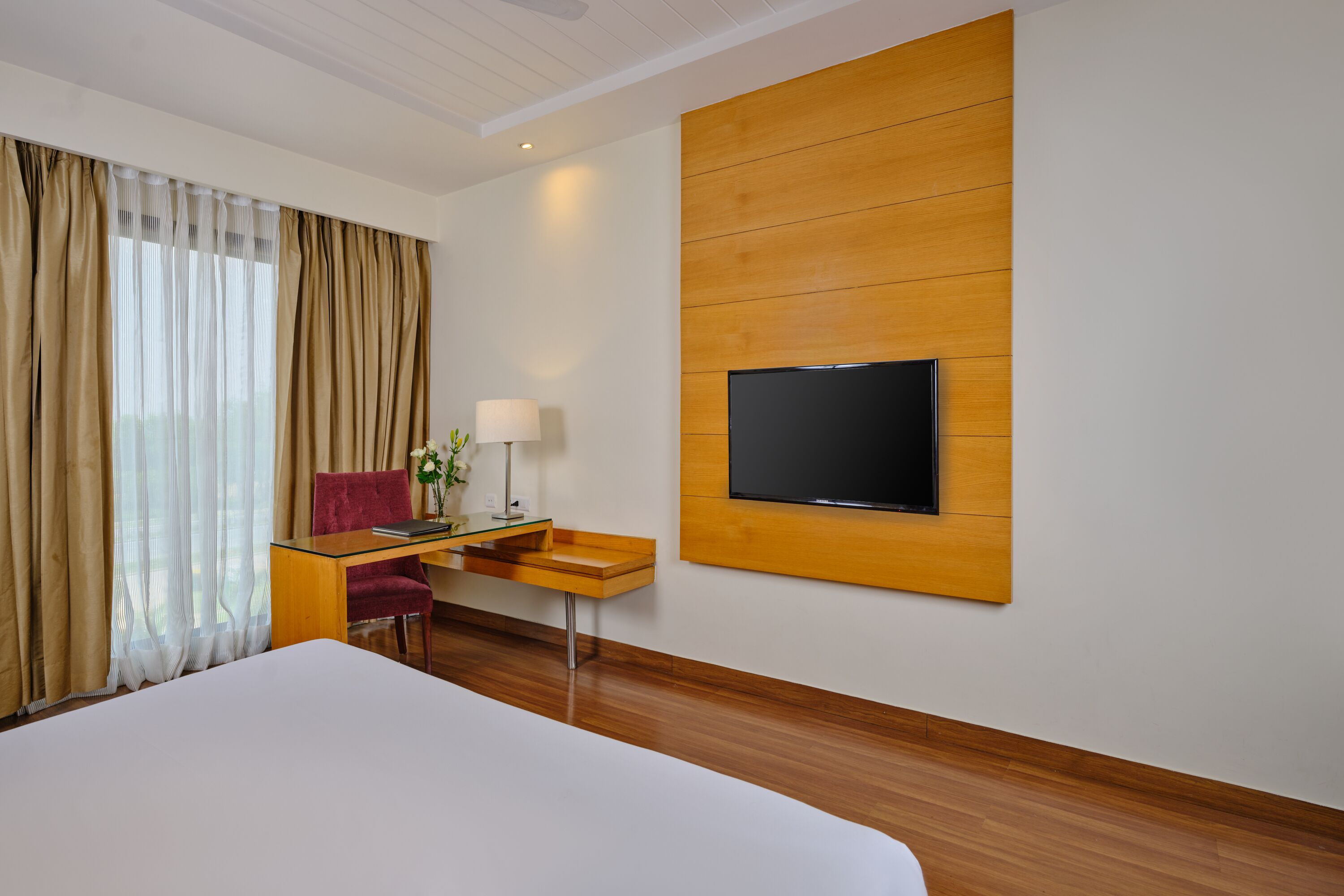 A hotel room with a large bed, a desk with a chair, a flat-screen TV mounted on the wall, and a window with curtains.