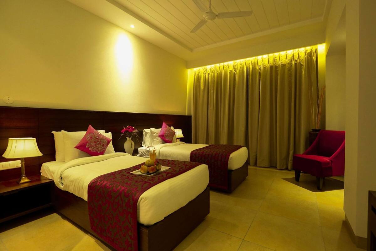 A hotel room with two beds, a nightstand, a lamp, a chair, and a large window with curtains.