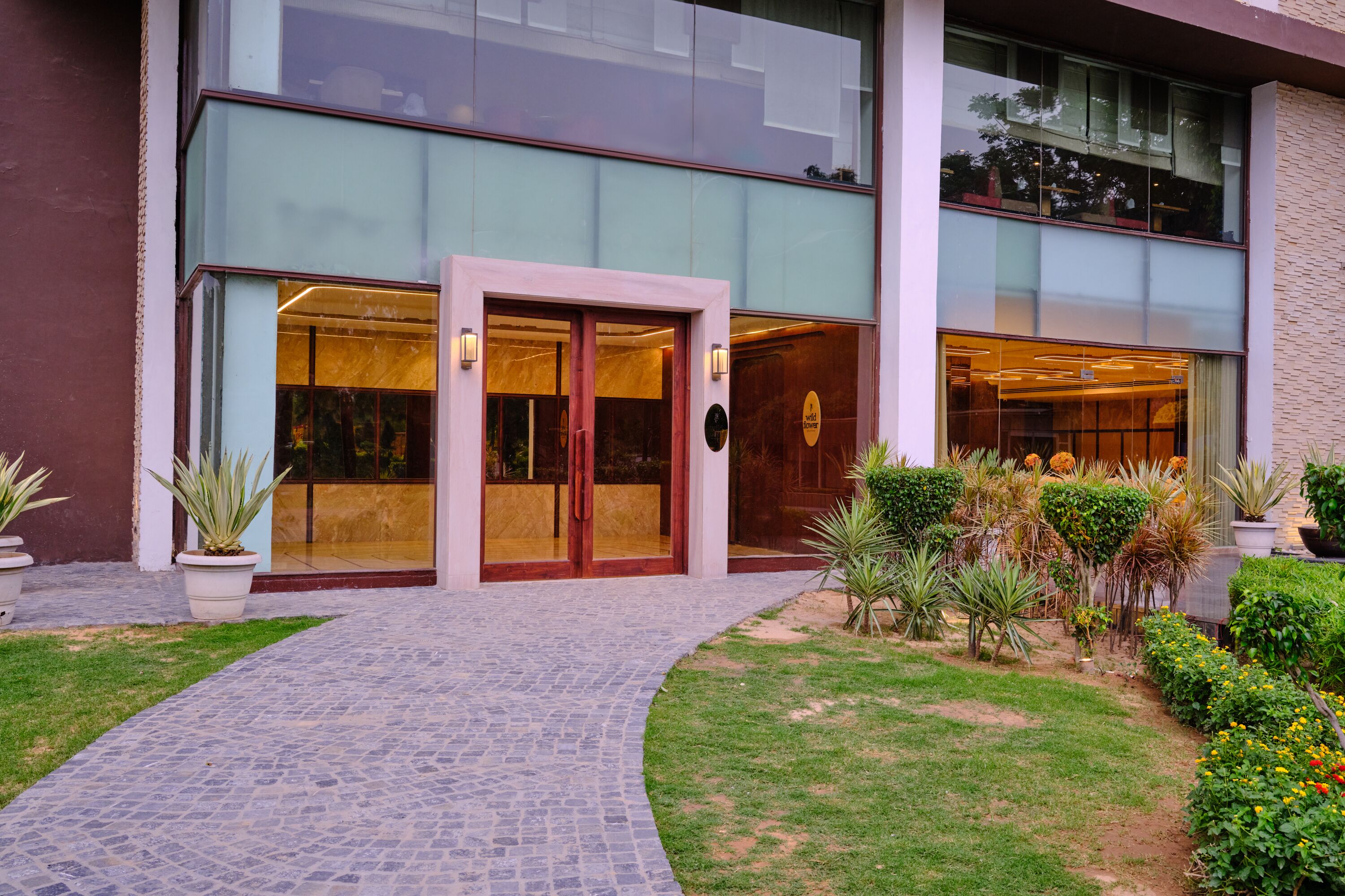 Un edificio moderno con entrada de vidrio, un camino pavimentado y un jardín bien cuidado con diversas plantas.