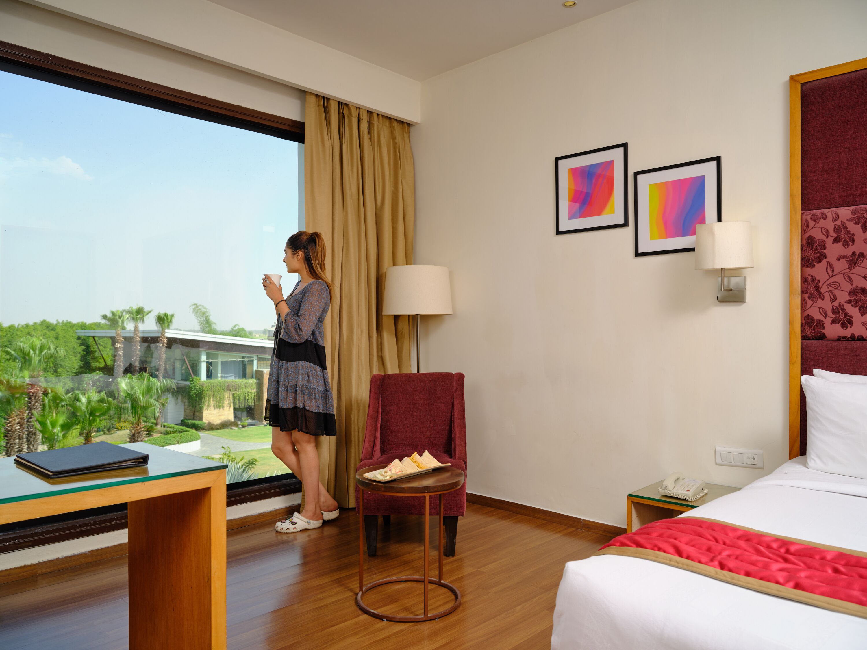 A hotel room with a large window, a bed, a desk, a chair, and a small table with a tray of food.