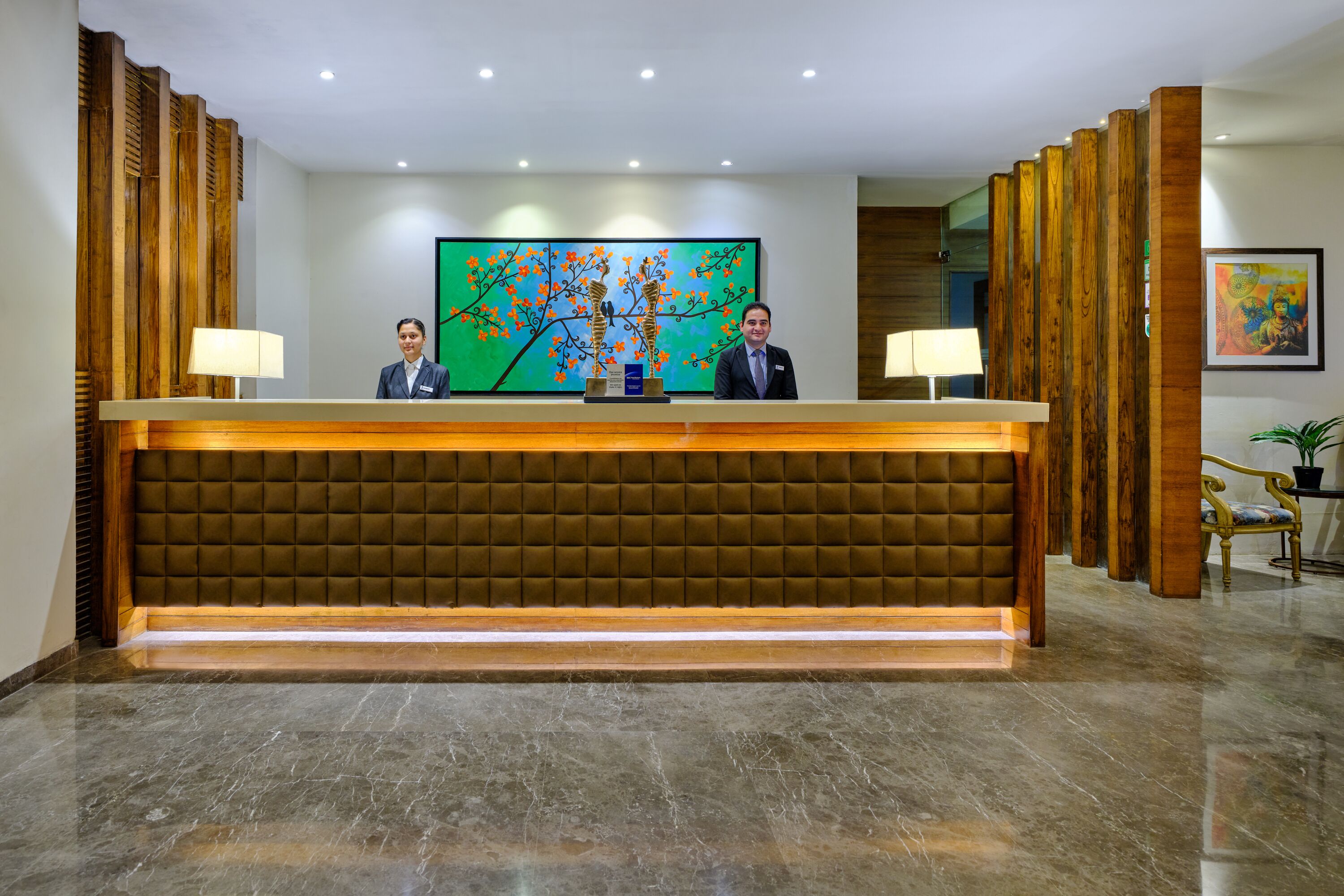 A reception area with a wooden reception desk, two receptionists, a green painting, and a potted plant.