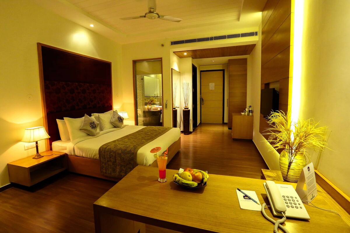 A hotel room with a bed, bedside table with a lamp, a fruit bowl, a telephone, and a vase with plants.