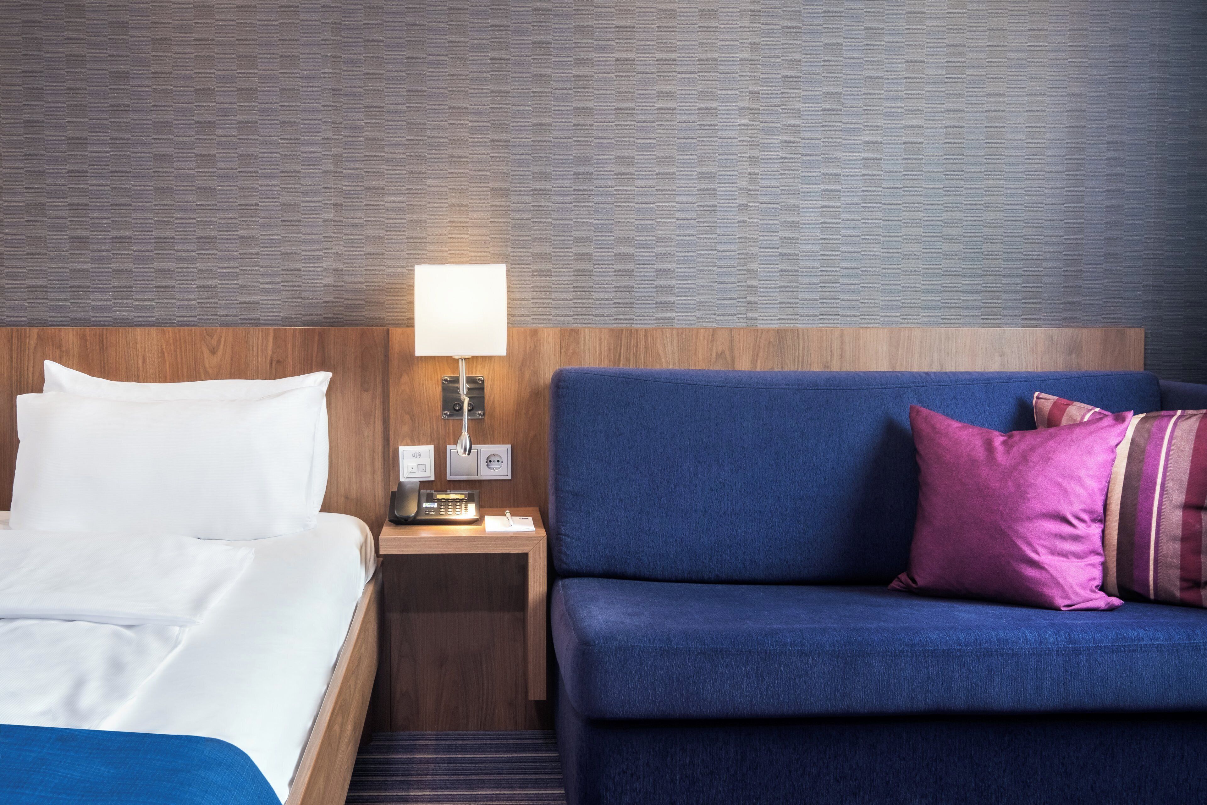 A hotel room with a bed, a nightstand with a lamp, and a sofa with pillows.