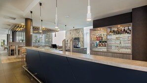 A modern bar area with a dark blue counter, hanging pendant lights, and a glass display of bottles and glasses. - Holiday Inn Express Friedrichshafen by IHG (Friedrichshafen)
