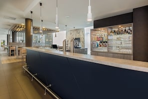 A modern bar area with a dark blue counter, hanging pendant lights, and a glass display of bottles and glasses. - Holiday Inn Express Friedrichshafen by IHG (Friedrichshafen)