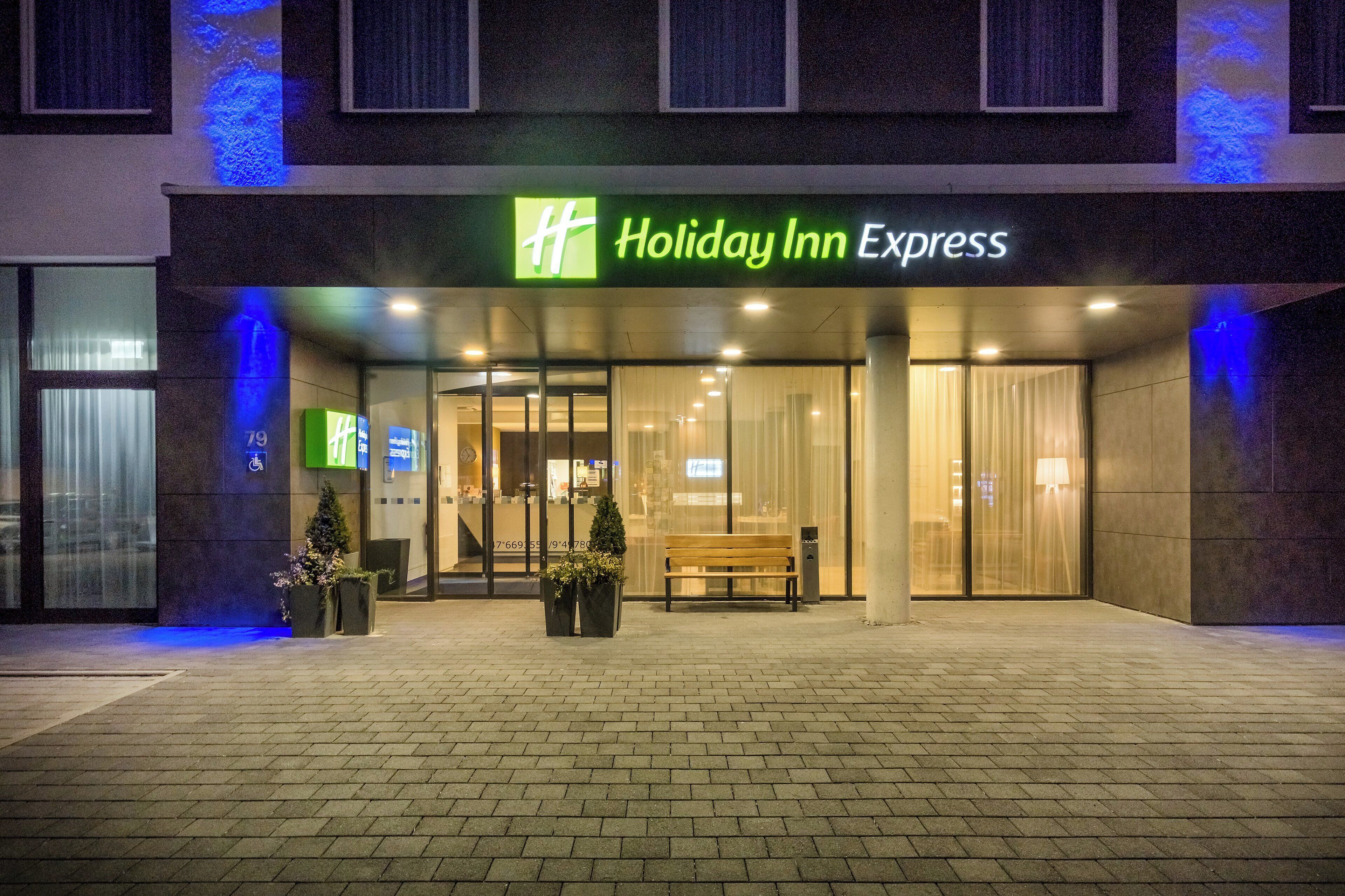 Edificio de Holiday Inn Express con entrada de vidrio, una banca en el exterior y plantas en macetas.