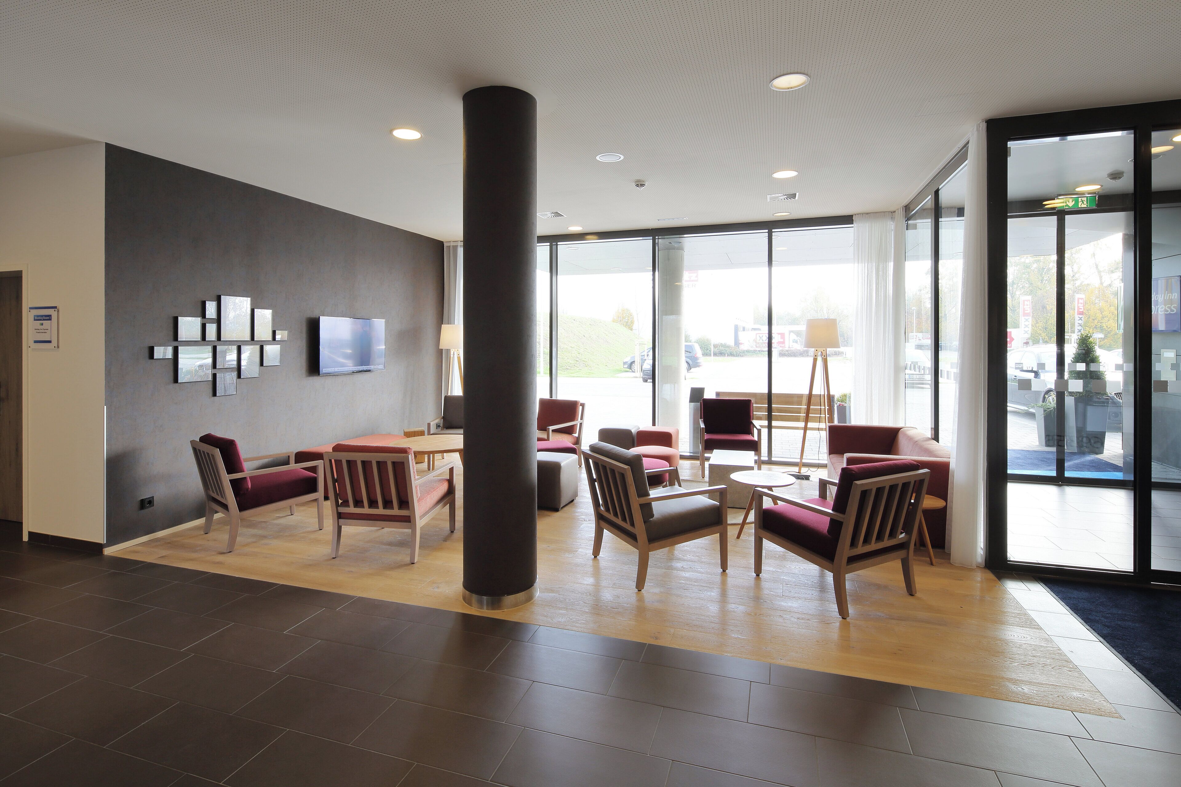 A spacious lobby with a large pillar, seating area with chairs and tables, a flat-screen TV mounted on the wall, and large windows offering a view of the outside.