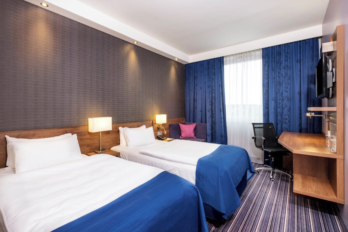 A hotel room with two beds, a desk, a chair, a TV, and a window with curtains.
