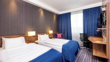 Ein Hotelzimmer mit zwei Betten, einem Schreibtisch, einem Stuhl, einem Fernseher und einem Fenster mit Vorhängen.