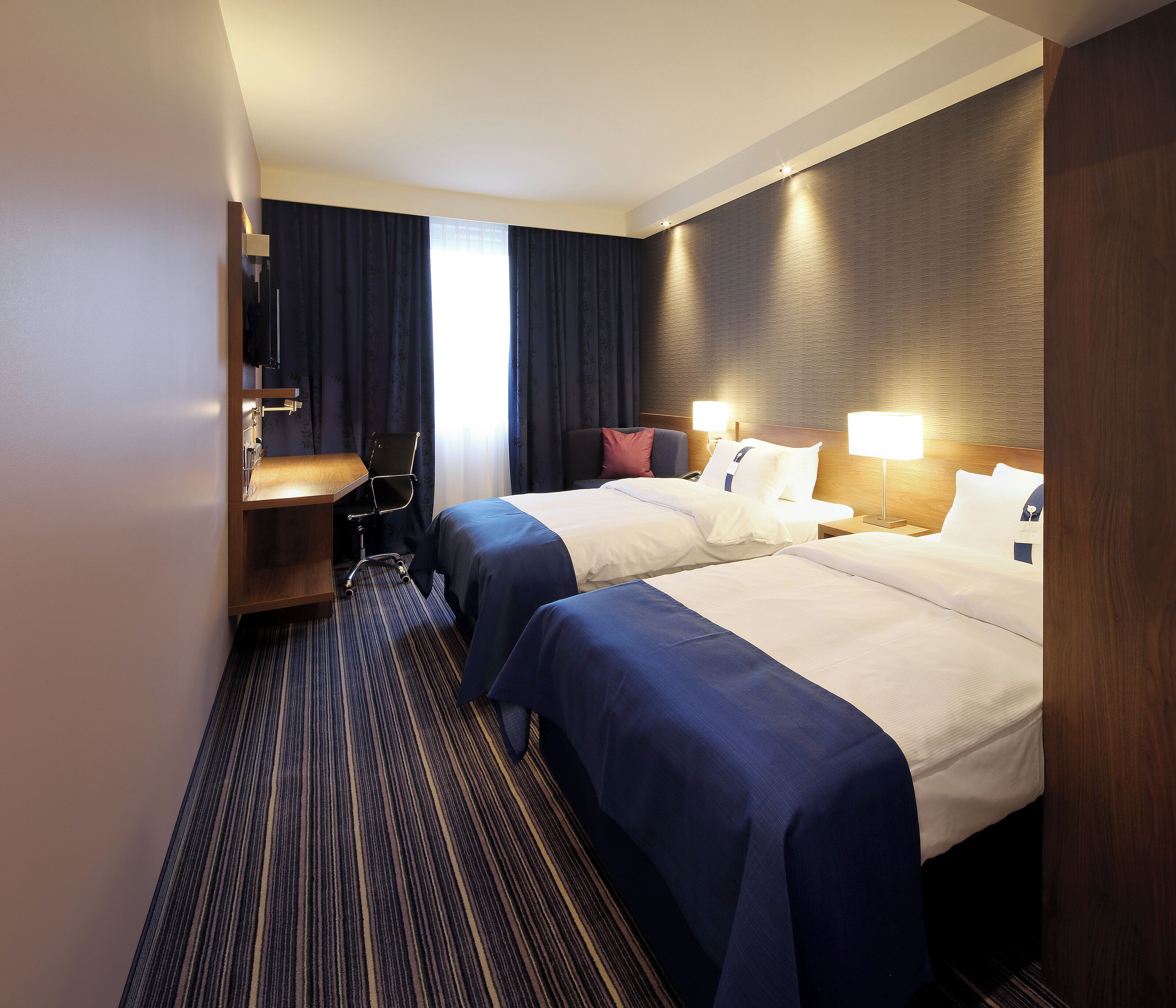 Ein Hotelzimmer mit zwei Betten, einem Schreibtisch, einem Stuhl und einem großen Fenster mit Vorhängen.