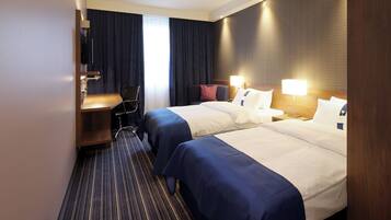 Ein Hotelzimmer mit zwei Betten, einem Schreibtisch, einem Stuhl und einem großen Fenster mit Vorhängen.