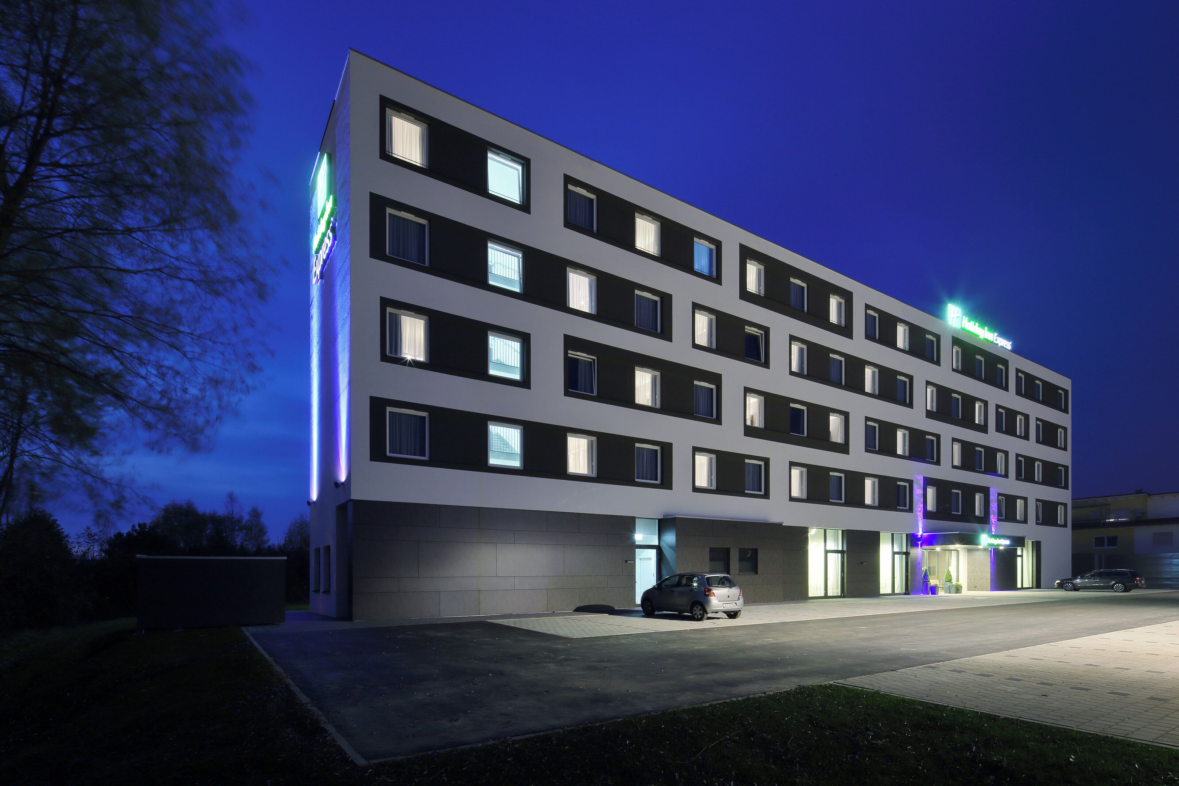 A multi-story hotel with a modern facade, multiple windows, and a visible entrance with a car parked outside.