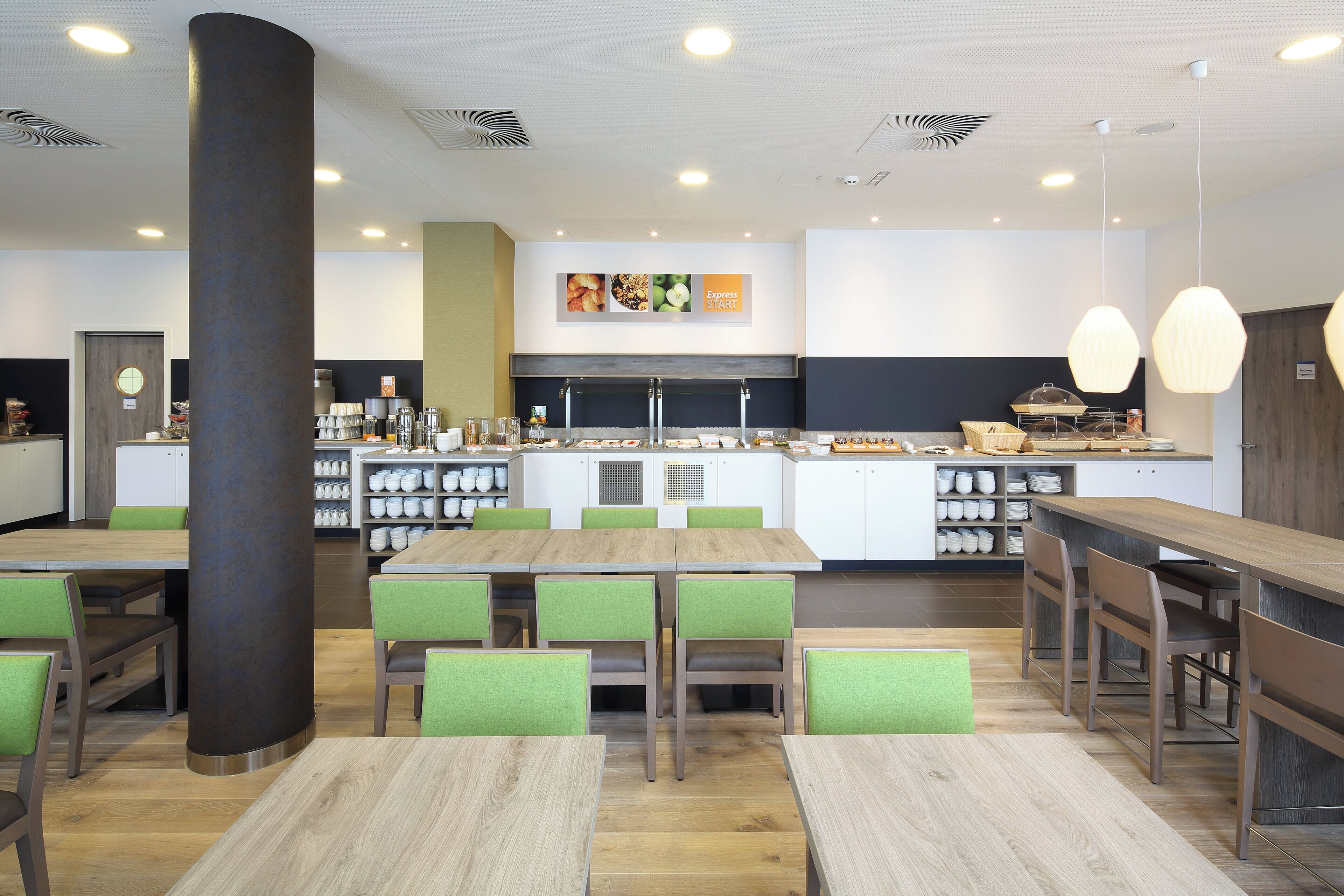 Een modern restaurant met een centraal eiland, groene stoelen met kussens en diverse serviesartikelen op planken.