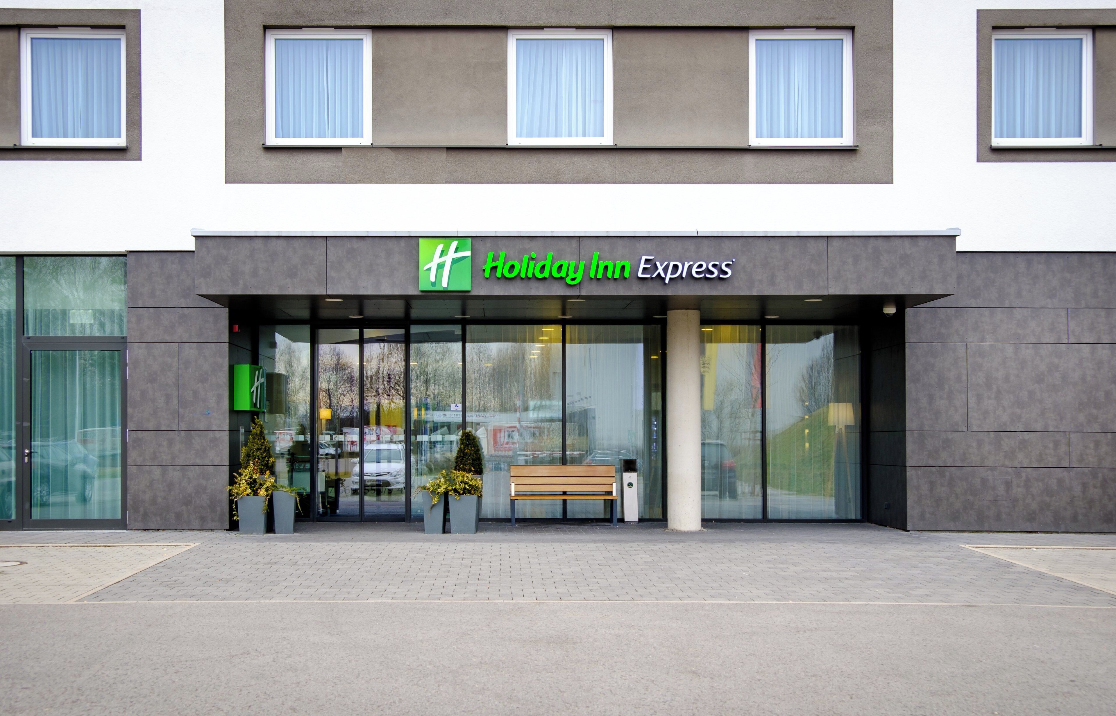 A Holiday Inn Express hotel entrance with a modern facade, large glass windows, and a bench outside.