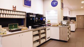 Um espaço com estação de café, várias máquinas de café, exposição de xícaras e diversas opções de comida.