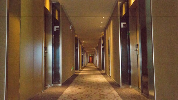 Een lange, smalle hotelgang met deuren aan beide kanten, een kamerbreed tapijt en een plafond met inbouwspots.