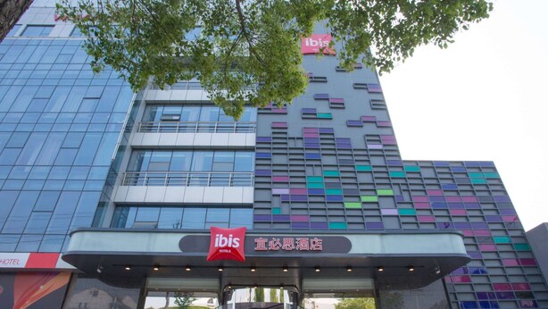 A hotel with a modern glass facade, a prominent red ibis logo, and a traditional Chinese sign above the entrance. - Ibis Changzhou Lihua Hotel (Changzhou)