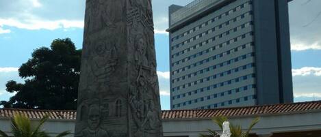 Un monumento alto con intrincados grabados, una fuente en primer plano y un edificio moderno de hotel al fondo.