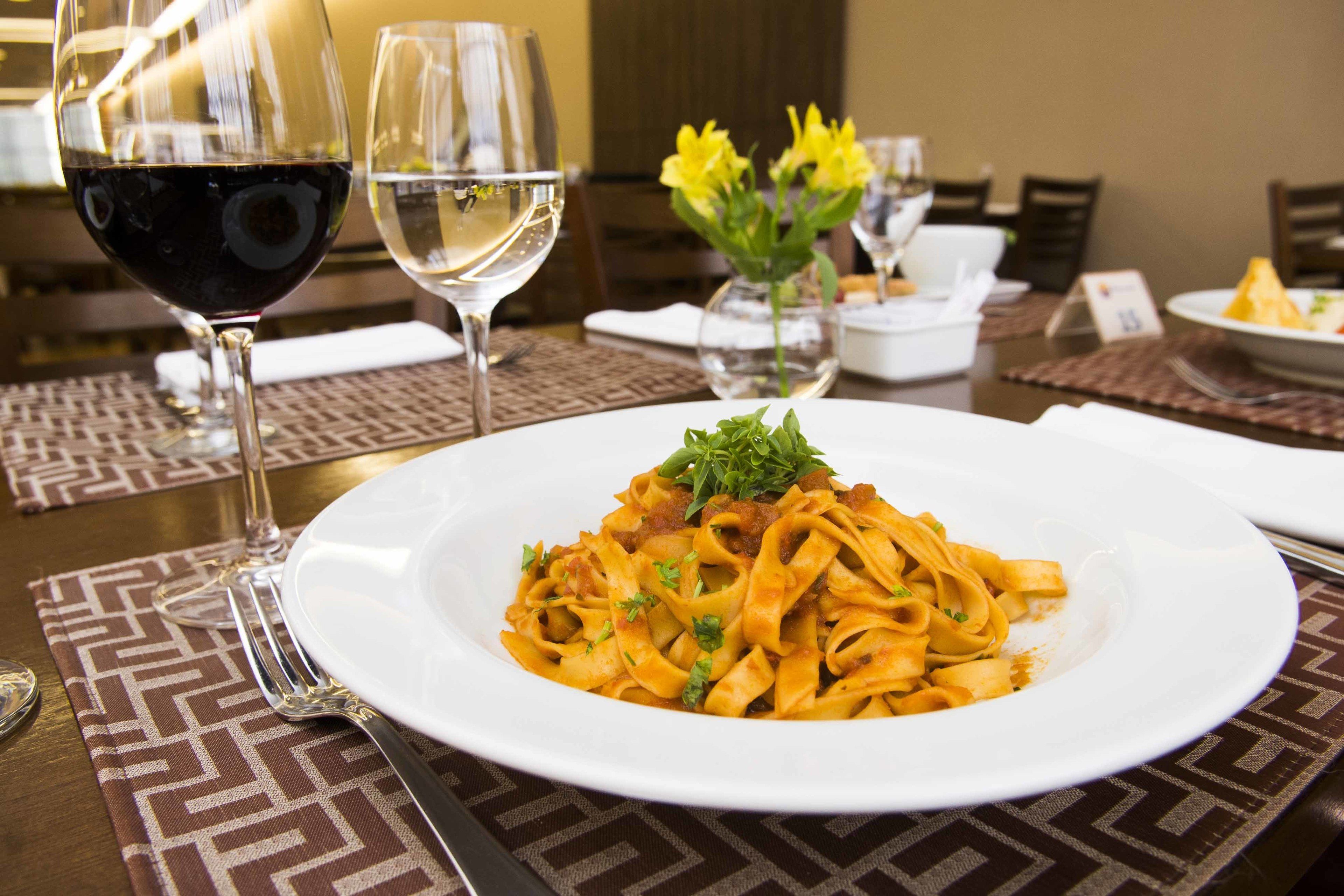 Una mesa de comedor con un plato de pasta, dos copas de vino y un florero con flores amarillas.