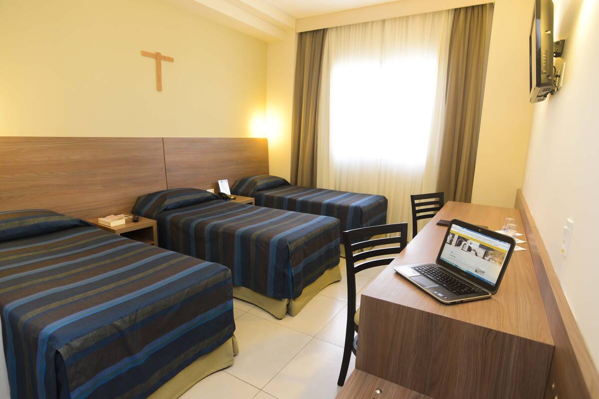 Een hotelkamer met twee eenpersoonsbedden, een bureau met een laptop, en een kruis aan de muur.
