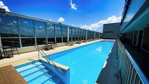 A large indoor swimming pool with clear blue water, surrounded by glass windows offering views of the sky and clouds. - Hotel Rainha do Brasil (Aparecida)