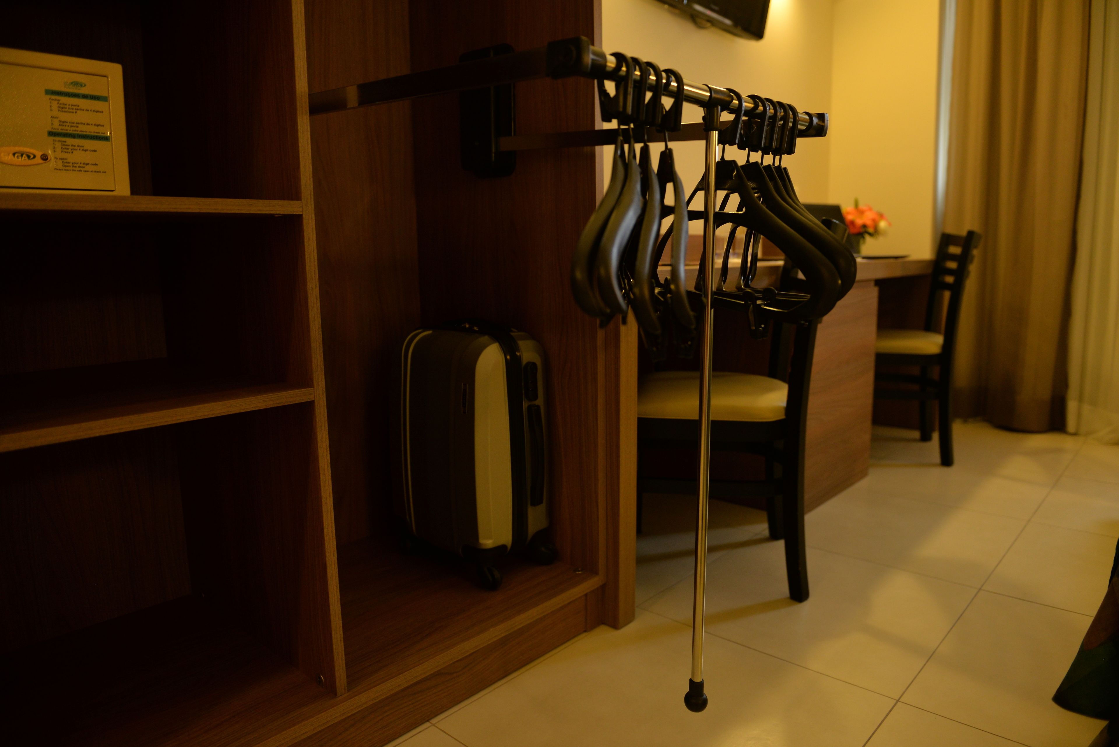 Une chambre d’hôtel avec une étagère en bois, une valise, un coin repas comprenant une table et des chaises, et un porte-manteau avec plusieurs vêtements.