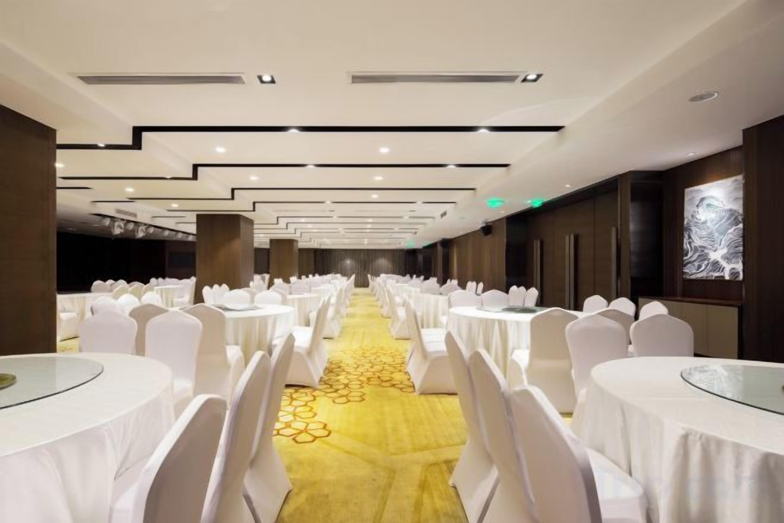 A banquet hall with rows of round tables, white chairs, and a yellow carpet.