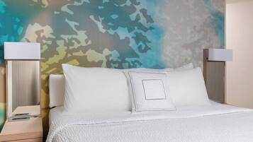 A neatly made bed with white bedding and a textured white throw blanket. There is a bedside table with a lamp and a drawer. The wall behind the bed features a large, abstract camouflage pattern.