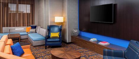 A modern hotel lobby with a sectional sofa, a blue armchair, a round coffee table, and a flat-screen TV mounted on the wall.