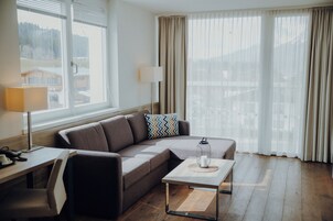 A modern living room with a sofa, a coffee table, and a lamp. There are large windows with curtains and a view of buildings outside.