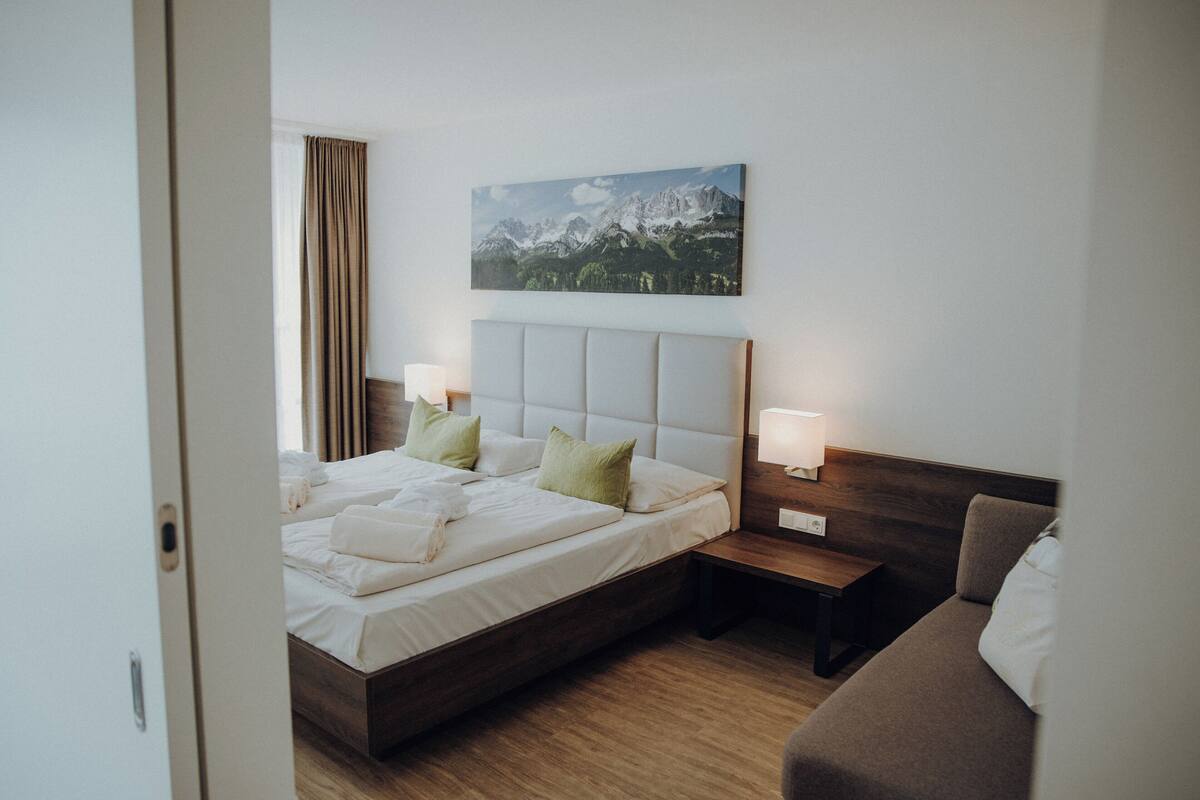 A hotel room with a bed, bedside tables, a nightstand, a sofa, and a painting on the wall.