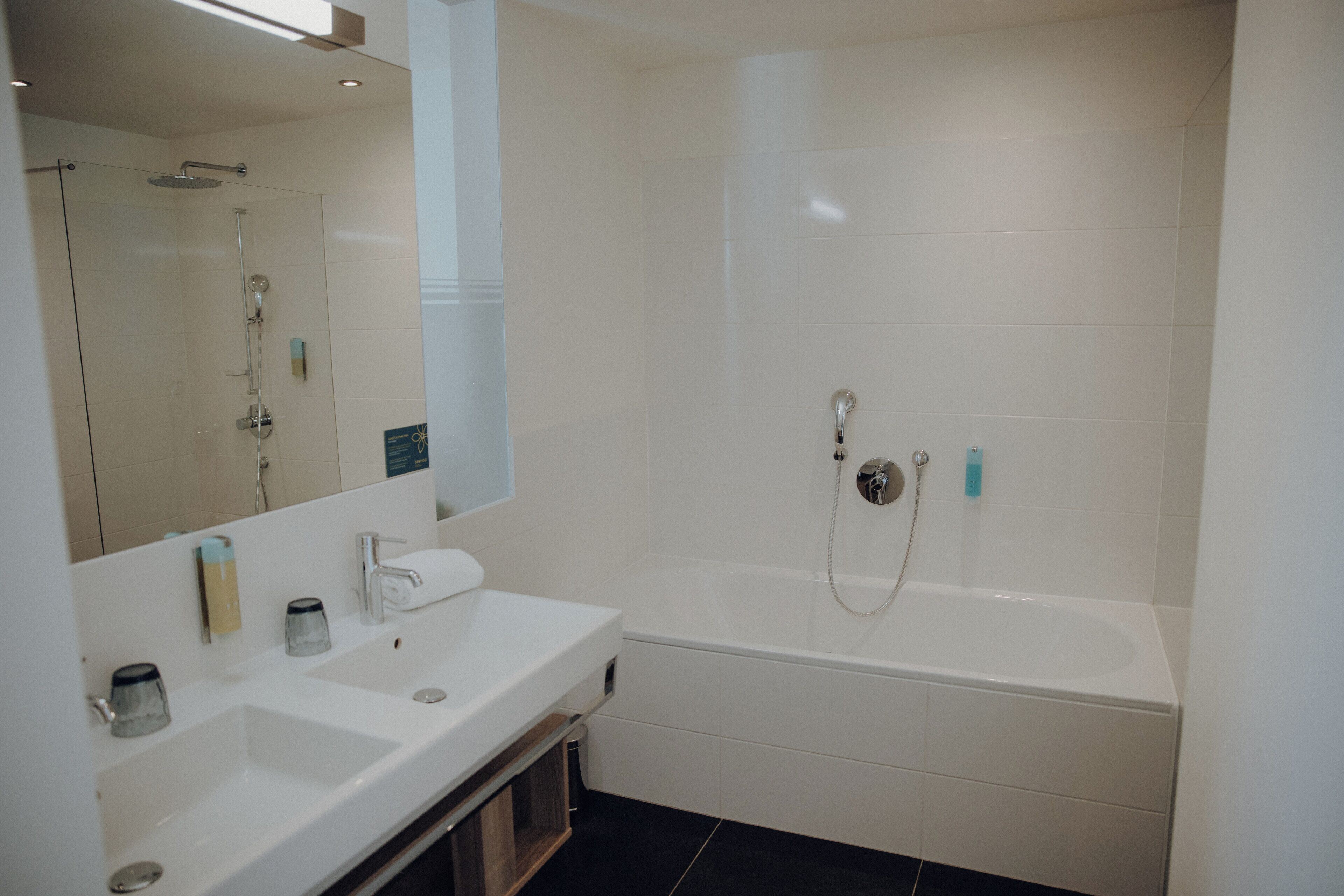A modern bathroom with a large mirror, two sinks, a bathtub, and a shower.