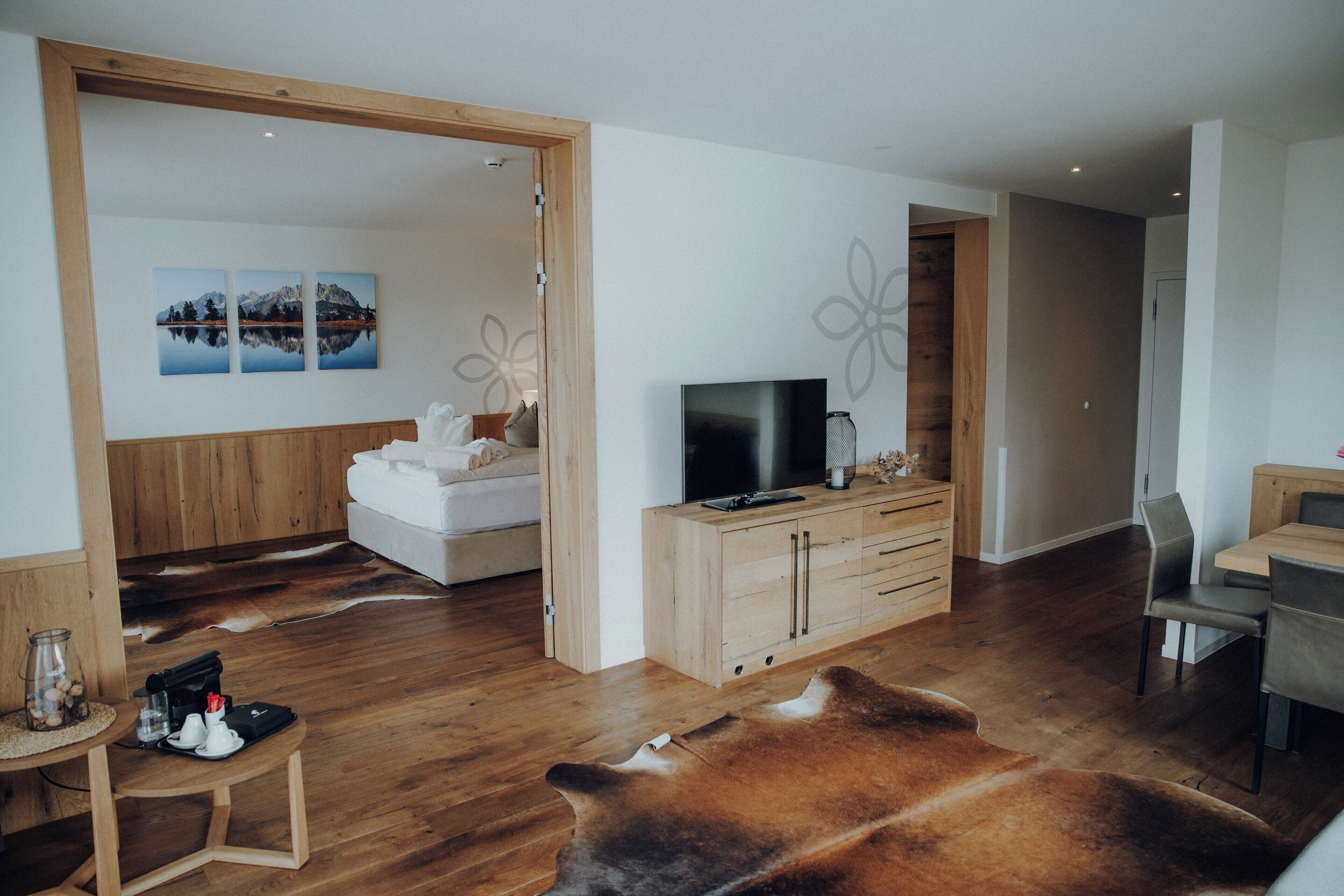 A modern living room with a wooden floor, a large leather sofa, a TV on a wooden cabinet, and a dining area with a table and chairs.