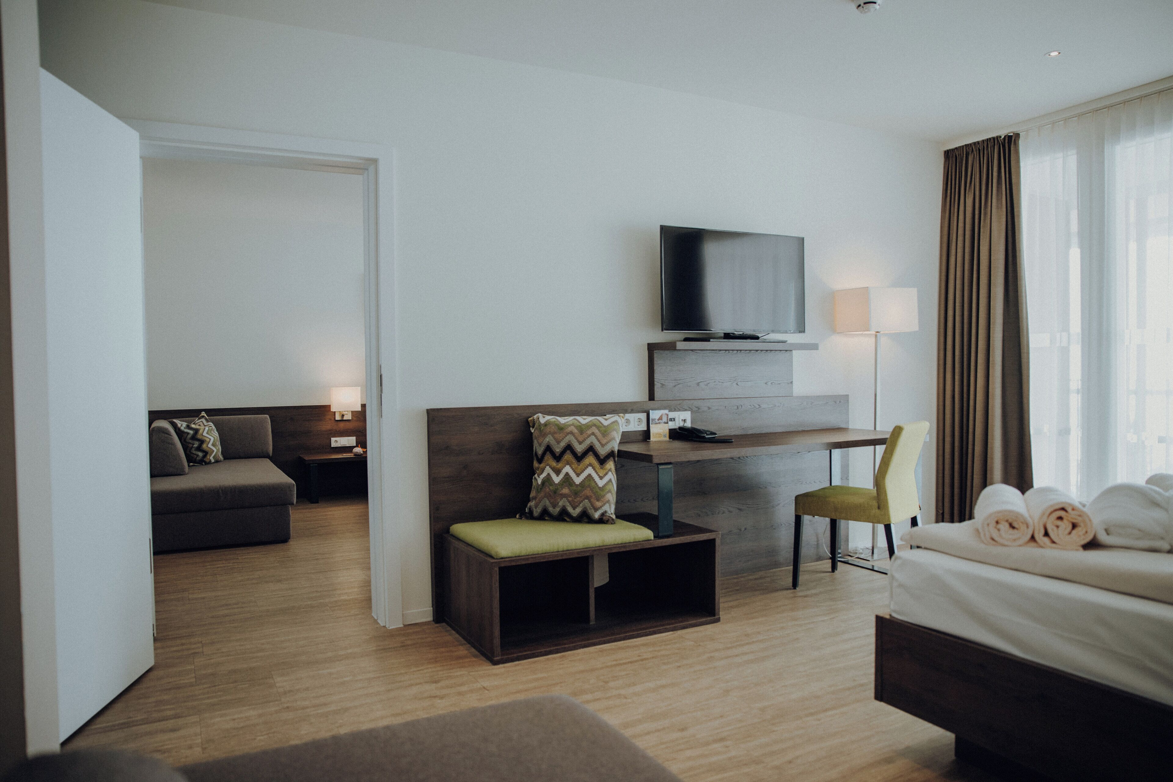 A hotel room with a bed, a desk with a TV, a chair, a sofa, and a lamp.