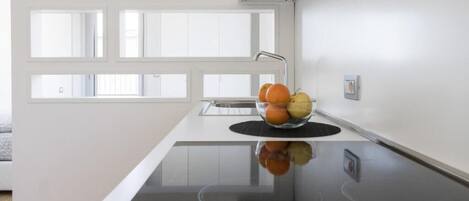 Een moderne keuken met een glazen kookplaat, een schaal fruit op het aanrecht en een raam met luxaflex.