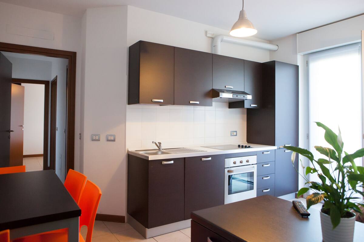 A modern kitchen with dark cabinets, a stainless steel sink, and a built-in oven. There is a dining table with orange chairs and a potted plant on the counter.