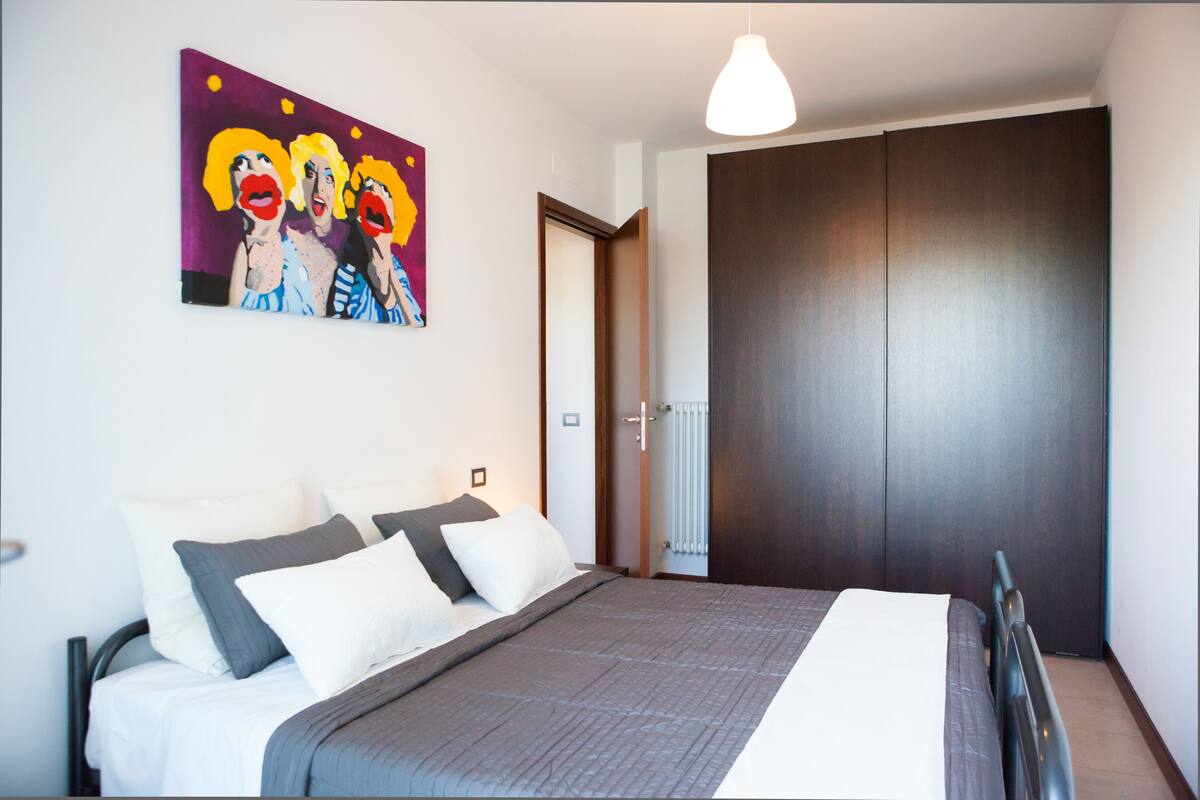 A bedroom with a bed, pillows, a painting, a wardrobe, and a door.