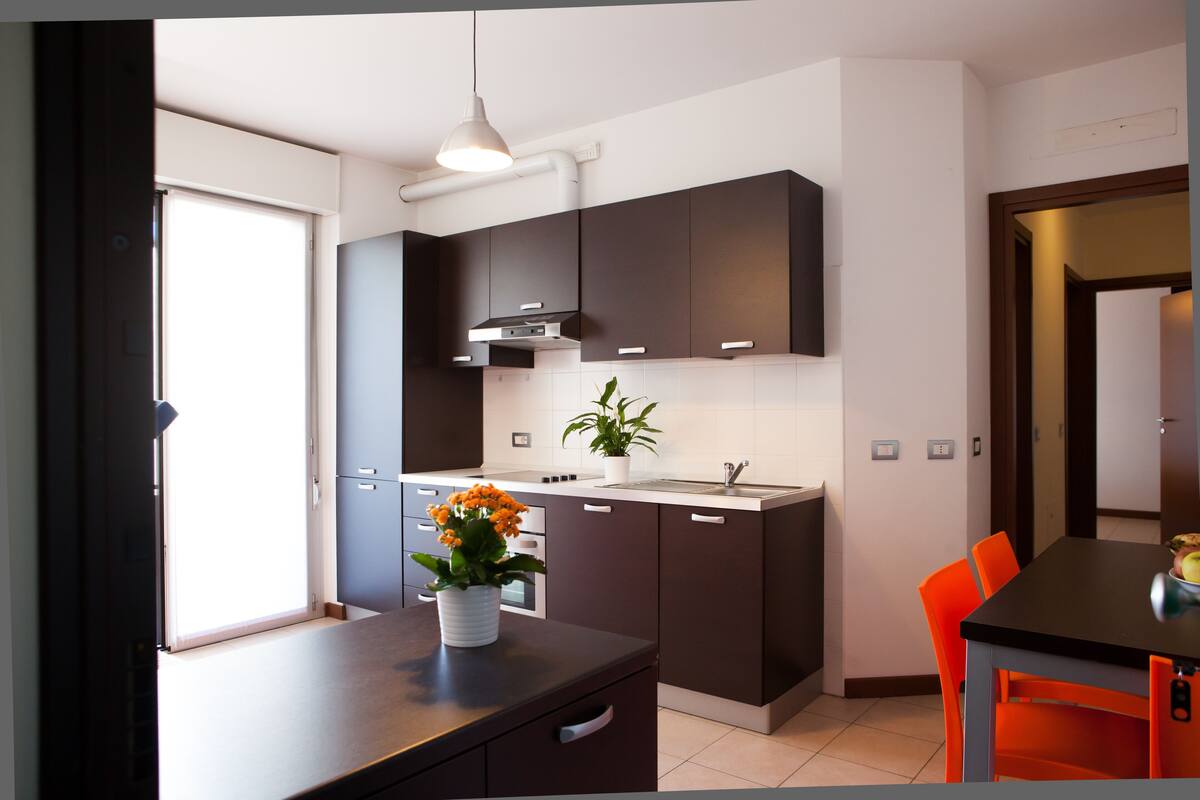A modern kitchen with dark cabinets, a white countertop, and a dining area with orange chairs.