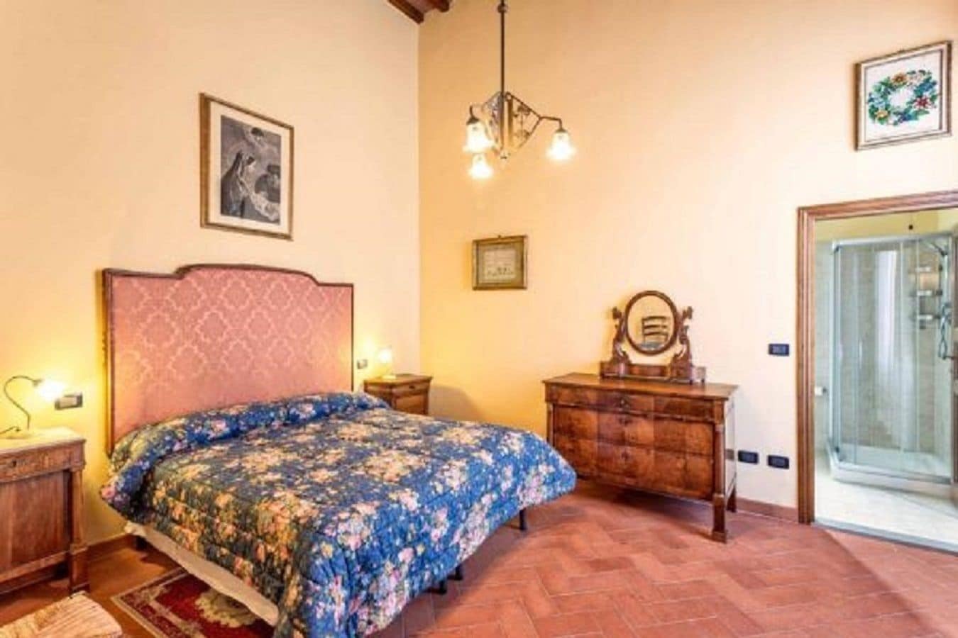 Un dormitorio con colcha estampada, cómoda de madera, espejo, lámpara de araña y zona de ducha.