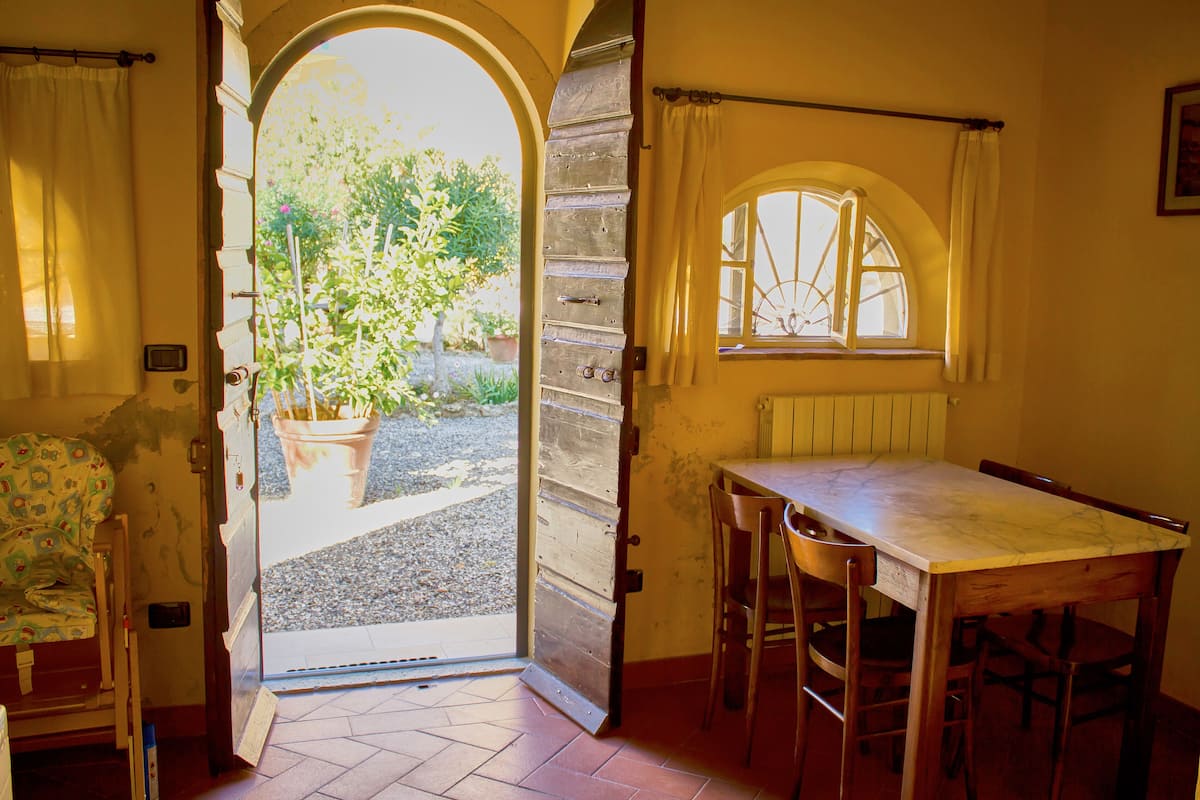 Eine offene Tür führt in einen Garten, ein Esstisch mit Stühlen und ein Fenster mit Vorhang.