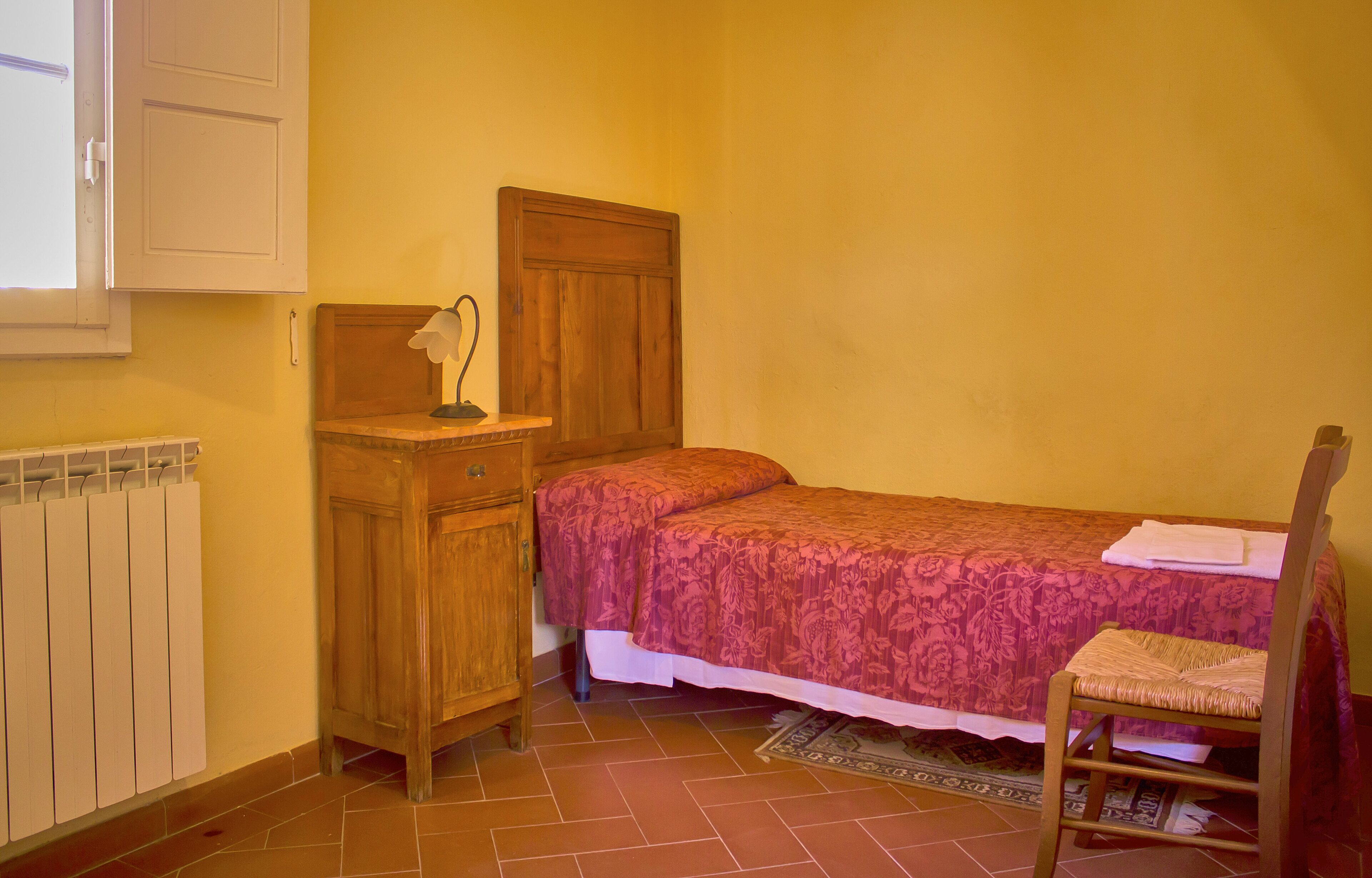 A small, neatly arranged bedroom with a single bed, a wooden nightstand, a chair, and a radiator.
