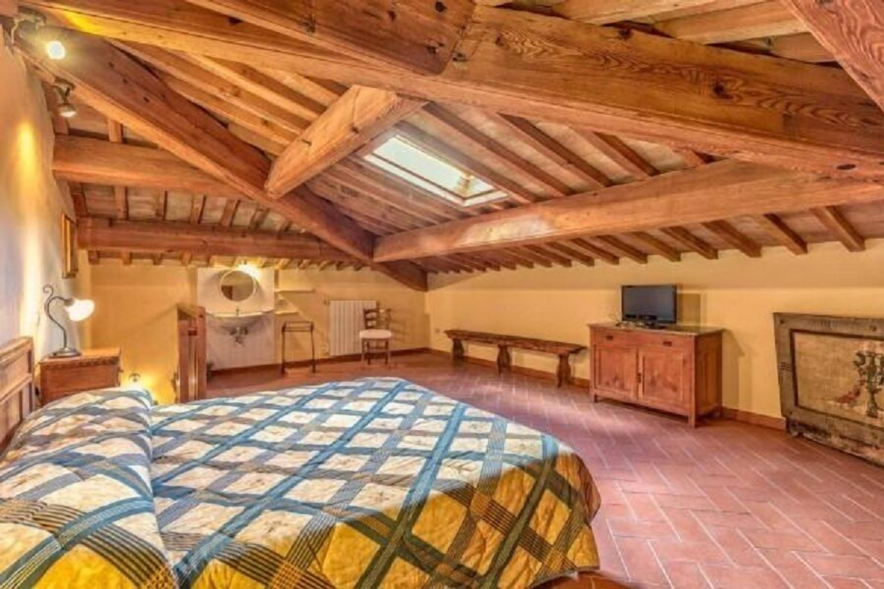 A bedroom with a wooden ceiling, a bed with a quilt, a dresser, a chair, and a television.