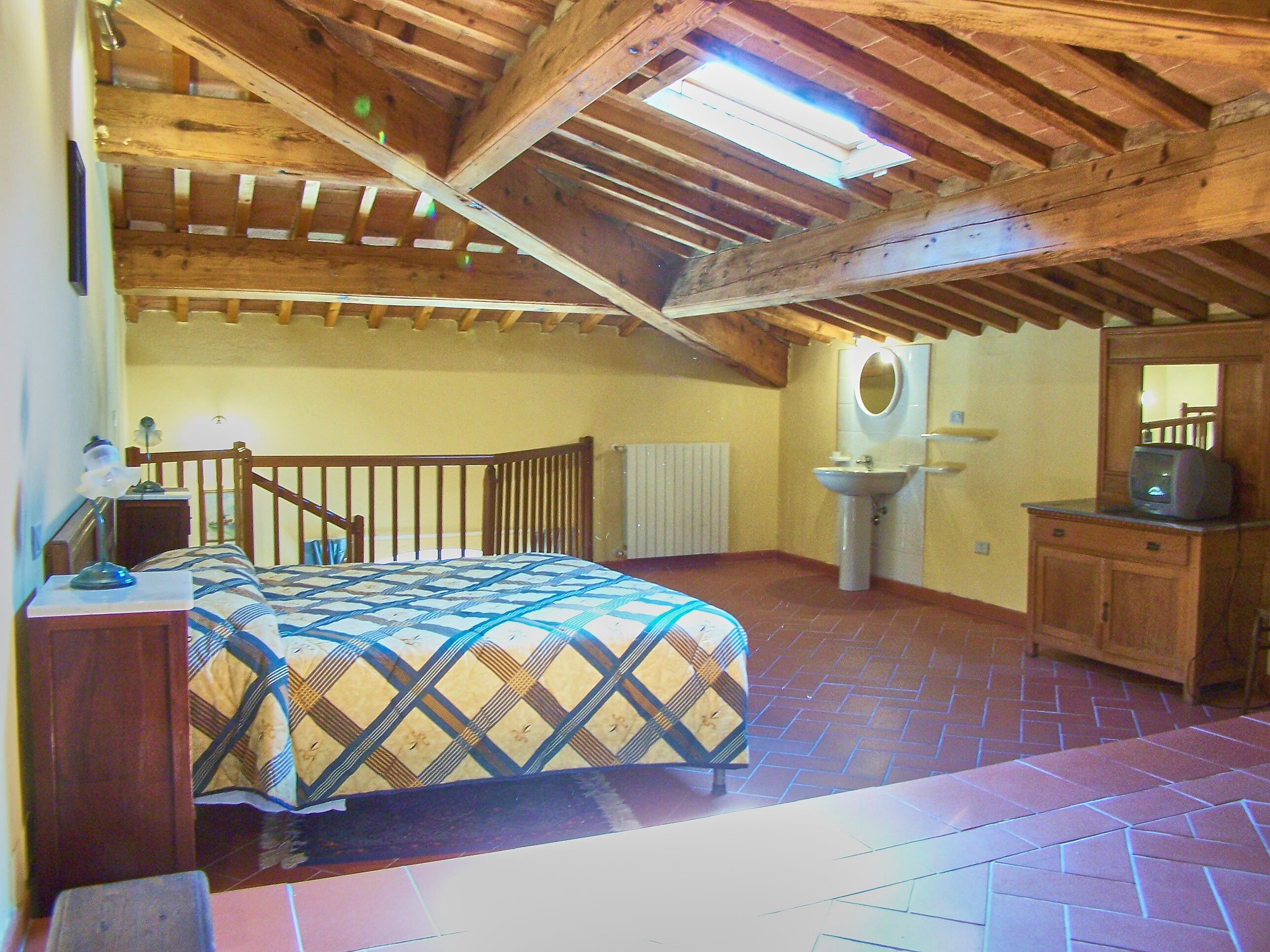 A bedroom with a wooden ceiling, a bed with a patterned bedspread, a bedside table with a lamp, a radiator, a sink, and a mirror.