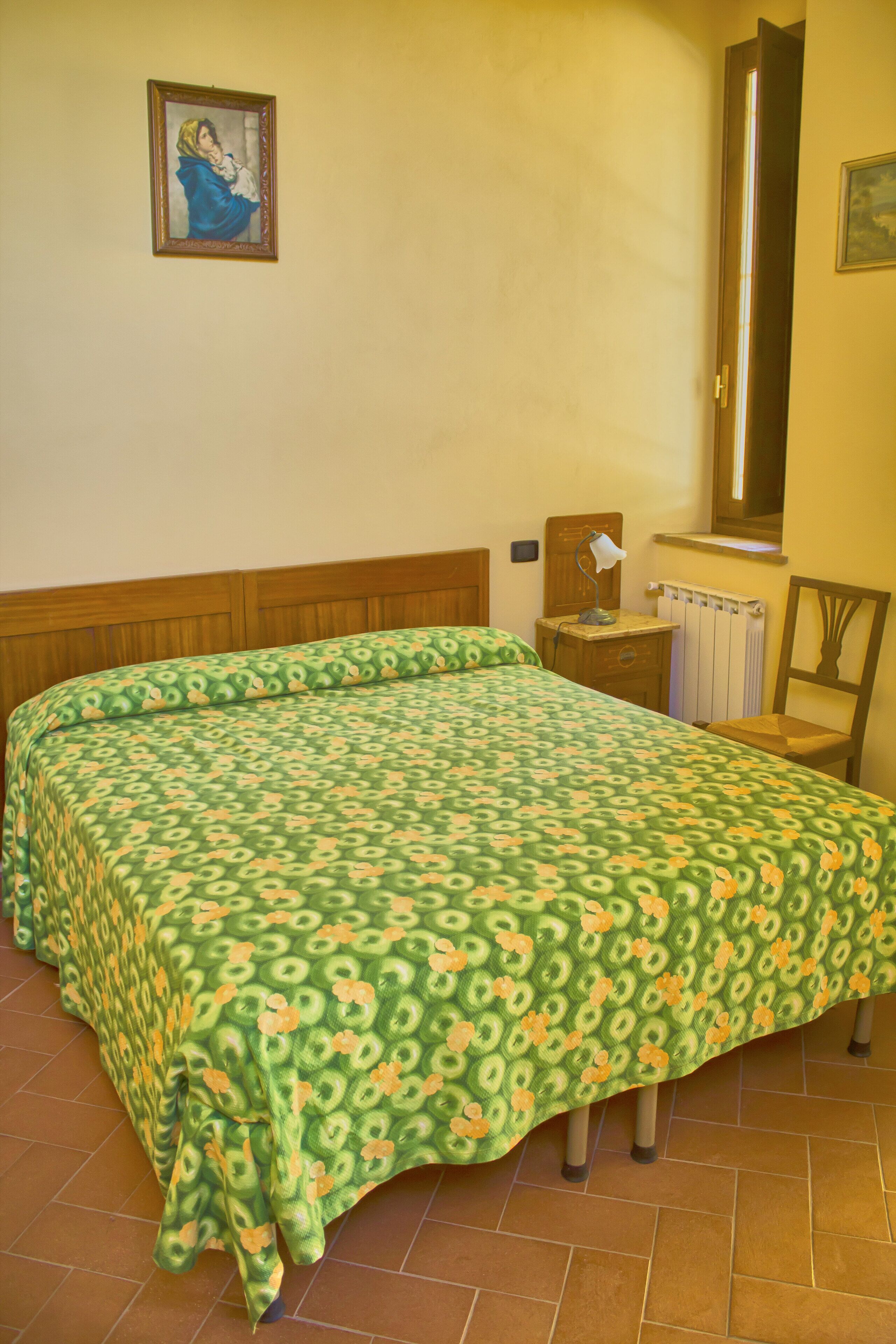A bedroom with a bed, a nightstand, a chair, a radiator, and a painting on the wall.