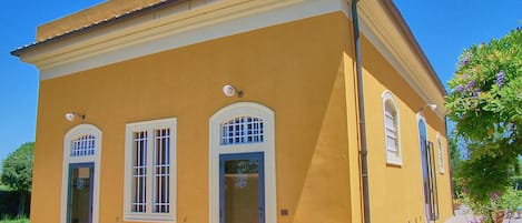 Ein gelbes Gebäude mit drei Rundbogenfenstern und einer Tür, umgeben von Kies und Topfpflanzen.