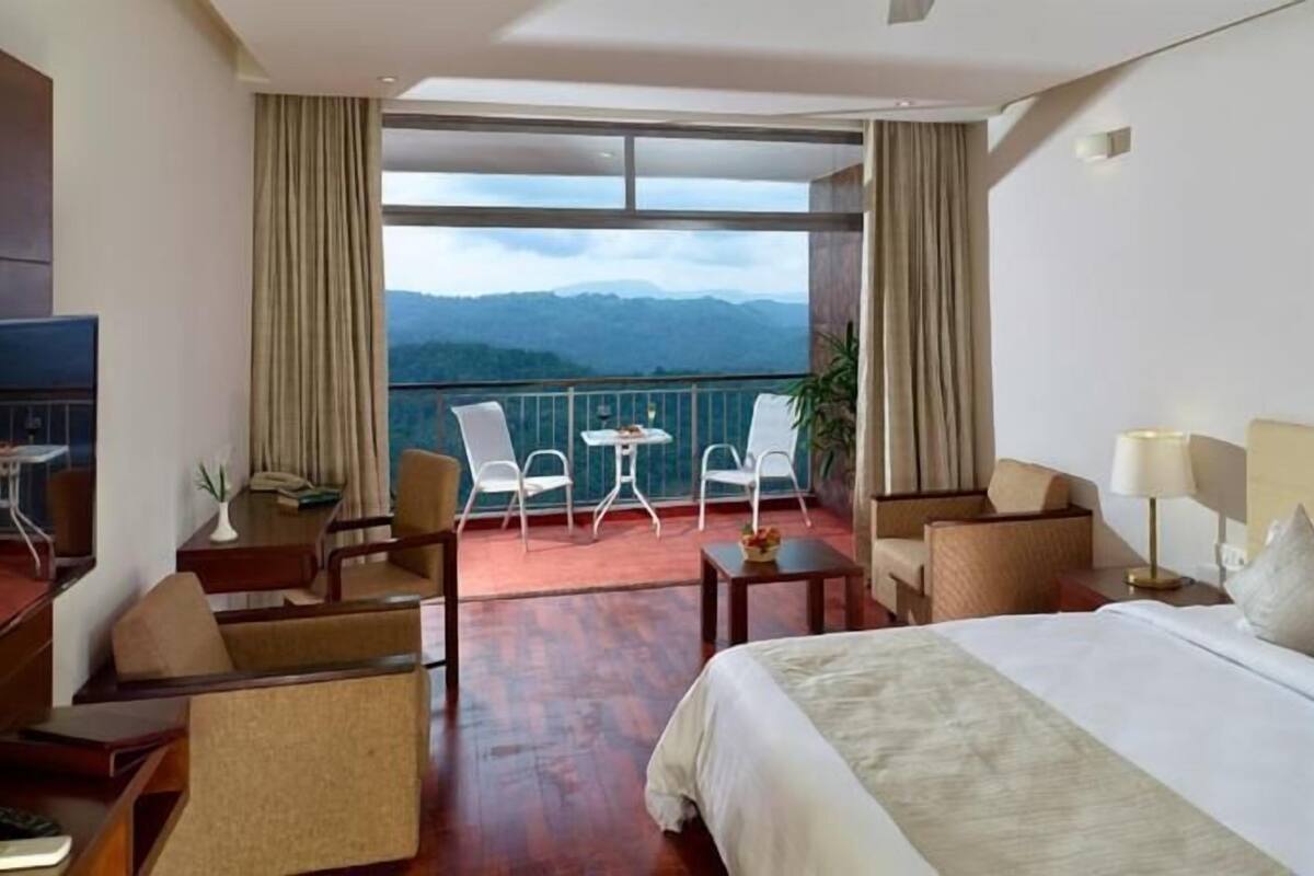A hotel room with a large bed, a balcony with chairs, a small table, and a painting on the wall.