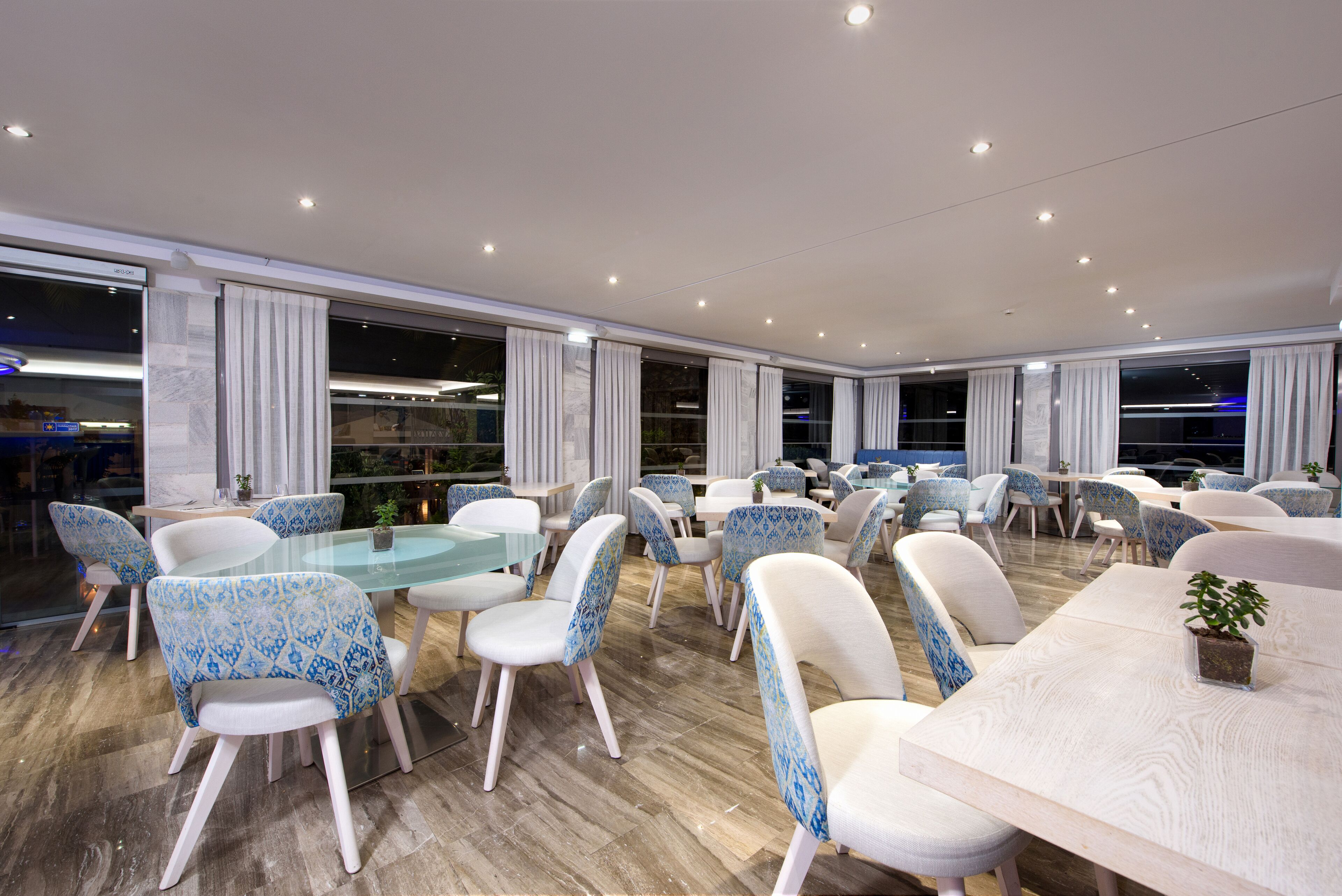 Un'ampia sala da pranzo con tavoli e sedie rotonde, grandi finestre con tende e un design d'interni moderno.